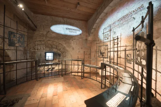Historische Museumsausstellung über Sündenböcke und Unterdrückung in einem Raum mit Steinwänden und Metallrahmen